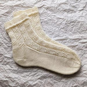 Pode incluir: Um par de meias brancas de malha com um padrão de renda. As meias são feitas de um material macio e confortável e têm um visual delicado e feminino.