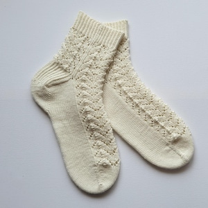 Könnte beinhalten: Ein Paar cremefarbene Strickstrümpfe mit einem detaillierten Lochmuster. Die Socken haben einen gerippten Bund und ein dekoratives Design, das die Vorderseite und den Knöchel hinunterläuft. Die Socken sind aus weichem, strukturiertem Garn gefertigt.