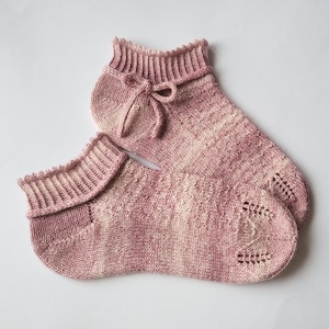 Könnte beinhalten: Ein Paar handgestrickte Knöchelsocken in Rosa- und Cremetönen. Die Socken haben einen gerippten Bund, eine dekorative Schleife und ein Muster auf der Oberseite des Fußes. Die Socken sind aus weichem Garn gefertigt.