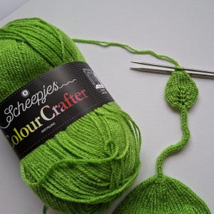 May include: A skein of bright lime green Scheepjes Colour Crafter yarn with a black label. A thin strand of the yarn extends from the skein to a pair of metal knitting needles, which are currently holding a small, knitted leaf-shaped piece.