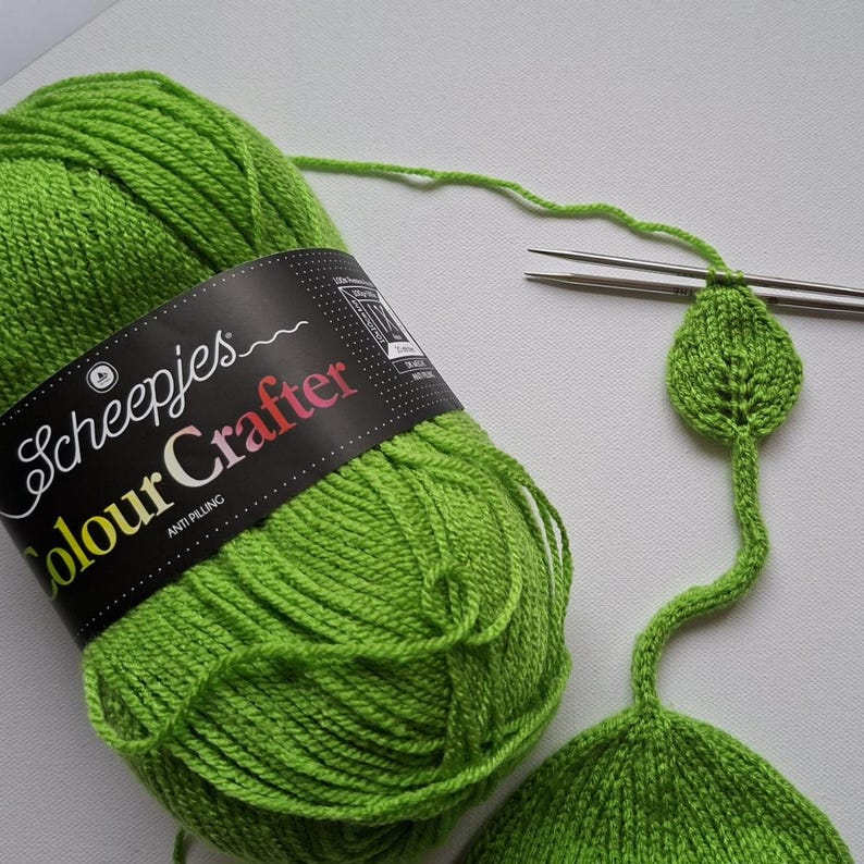 May include: A skein of bright lime green Scheepjes Colour Crafter yarn with a black label. A thin strand of the yarn extends from the skein to a pair of metal knitting needles, which are currently holding a small, knitted leaf-shaped piece.