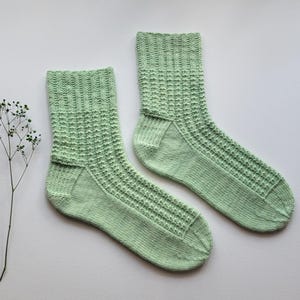 Può includere: Un paio di calzini lavorati a maglia color verde chiaro con un motivo a nido d'ape testurizzato sulla gamba e sul dorso del piede, dotati di un polsino a coste e una lavorazione a maglia liscia su tallone, suola e punta. Un piccolo stelo di fiore secco è posto accanto.
