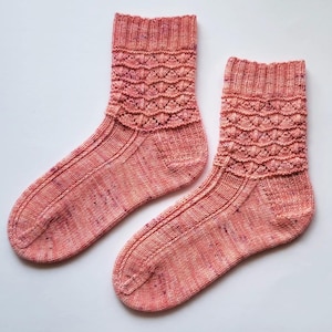 Könnte beinhalten: Ein Paar handgestrickte Socken in Rosa- und Pfirsichtönen. Die Socken haben ein strukturiertes, wellenförmiges Muster am Bündchen und ein gesprenkeltes Design am ganzen Körper. Die Socken werden vor einem weißen Hintergrund präsentiert.