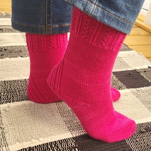 Può includere: Un paio di calzini rosa acceso a maglia con un motivo strutturato nella parte superiore e un design a coste sulla gamba. I calzini sono indossati con jeans di denim blu. I calzini sono su un tappeto a strisce bianche e nere.