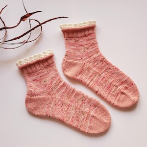 May include: A pair of pink knit socks with a white, scalloped trim at the top. The socks have a speckled pattern with hints of red, green, and yellow. The socks are displayed on a white surface.