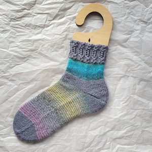 Puede incluir: Un calcetín a rayas gris, azul y amarillo con un patrón de encaje en la parte superior, colgado de una percha de madera.