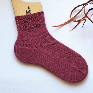 May include: A single burgundy knitted sock on a wooden form. The sock has an openwork pattern at the cuff and a ribbed texture on the leg and foot. The background is white, with a dried plant stem adding a natural accent.