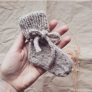 May include: A beige knitted baby bootie with a tied bow. The bootie is made of a soft, textured yarn.