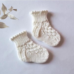Peut inclure: Une paire de chaussettes de bébé blanc cassé. Les chaussettes sont tricotées avec un motif texturé sur le pied et un bord-côte. Les poignets ont un bord festonné. Les chaussettes sont en matière douce, possiblement en laine.