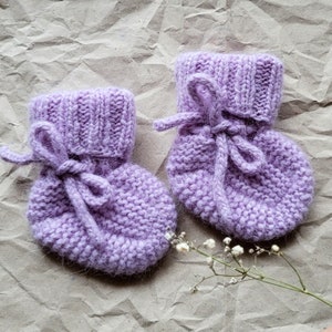 Pode incluir: Um par de botinhas de bebê de tricô lavanda com um laço amarrado em cada uma. As botinhas estão sobre um fundo de papel marrom amassado.
