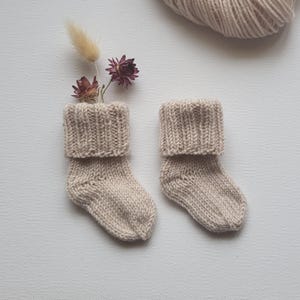 May include: Two beige knitted baby socks with folded cuffs. The socks are displayed with dried flowers and a ball of yarn in the background. The socks are made of a soft, textured yarn, perfect for a newborn.
