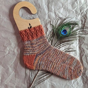 Op de afbeelding: Een paar handgebreide sokken van multicolor garen met oranje, bruin en paarse tinten. De sokken hebben een decoratief kabelpatroon aan de bovenkant en worden gepresenteerd op een houten hanger.