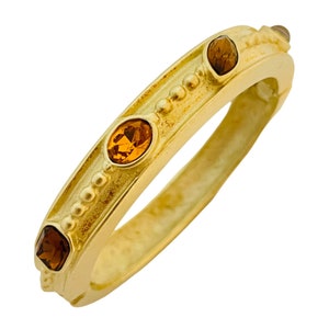 Puede incluir: Un brazalete dorado con tres piedras preciosas de color ámbar engastadas en la banda. El brazalete tiene un diseño de cuentas alrededor del borde.