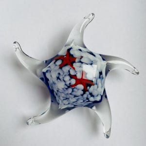 May include: Glass starfish sculpture. The clear glass has red starfish accents and a white and blue speckled interior. The sculpture has five pointed arms and is a decorative item.