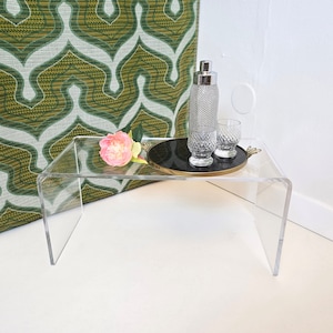 Puede incluir: Una mesa de acrílico transparente con una bandeja negra y ribete dorado. Una flor rosa, dos vasos transparentes y una coctelera de plata están en la bandeja.