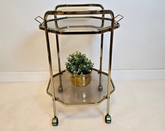 Puede incluir: Un carrito de servicio de dos niveles hecho de metal dorado con un marco octogonal y estantes de vidrio transparente. El carrito cuenta con cuatro ruedas pequeñas en la base y dos asas laterales. Una pequeña planta verde en maceta se encuentra en el estante inferior.