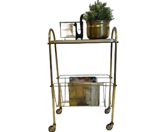 May include: A two-tiered gold-toned metal rolling trolley with a top shelf and a lower wire magazine rack. The top shelf holds a metallic planter with a green plant, a black abstract sculpture, and a rectangular frame. The trolley has four wheels.