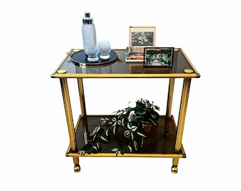Puede incluir: Un carrito de dos niveles con estructura de metal dorado y estantes de cristal oscuro. El estante superior contiene una coctelera plateada, dos vasos texturizados sobre una bandeja redonda y dos marcos. Una planta verde se encuentra en el estante inferior.