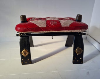 May include: A rectangular stool with a padded seat featuring a red and beige patchwork pattern. The base is constructed from black wood with four legs, each accented with circular gold-toned metal plates and small brass studs at the bottom.