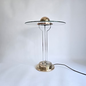 Può includere: Una lampada da tavolo in cromo e ottone con un paralume in vetro e una parte superiore rotonda a forma di cupola. La lampada ha una base a tre gambe e un cavo nero.