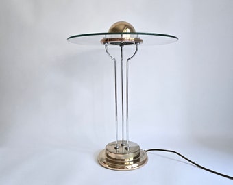 May include: A table lamp featuring a circular clear glass shade topped with a polished brass dome. The lamp has three slender chrome vertical supports rising from a tiered, circular brass base. A thin black power cord extends from the base.