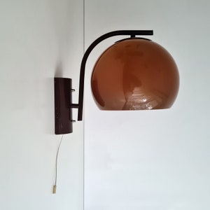 Puede incluir: Una lámpara de pared con una pantalla esférica marrón y un brazo de metal marrón oscuro. La lámpara está fijada a la pared con una base marrón oscuro a juego. Un cordón cuelga debajo.