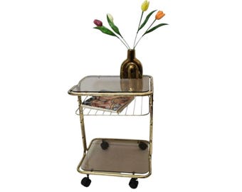 May include: A two-tier rolling cart with a gold-toned metal frame and four black casters. It features two square shelves made of smoked glass. A gold-toned vase containing four artificial tulips in yellow, white, purple, and orange sits on top.