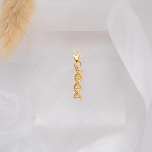 May include: A gold-colored DNA charm with a lobster clasp. The charm is a detailed representation of a DNA double helix. The background is a soft white fabric.