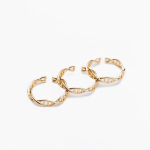 May include: Three gold-coloured rings, each with a DNA double helix design. The rings are open-ended for adjustable sizing. The jewellery is set against a plain white background, highlighting the intricate details of the rings.