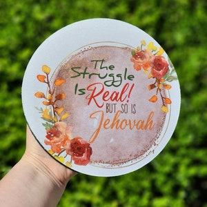 May include: A round white coaster with a brown and gold circle in the center. The circle has a watercolor design of flowers and leaves in shades of orange, red, and yellow. The text "The Struggle Is Real! But So Is Jehovah" is written in black and gold lettering.