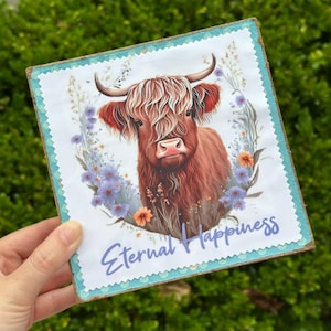 May include: A square wooden sign featuring a watercolor illustration of a Highland cow with long, shaggy red-brown fur, surrounded by a floral wreath. The sign has the text "Eternal Happiness" in a script font.