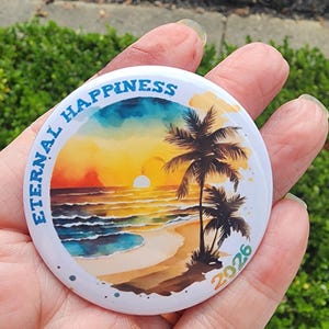 May include: A round button with a beach scene featuring palm trees, a sunset, and the words "ETERNAL HAPPINESS" in blue. The year "2026" is printed in yellow. The button is held in a hand.