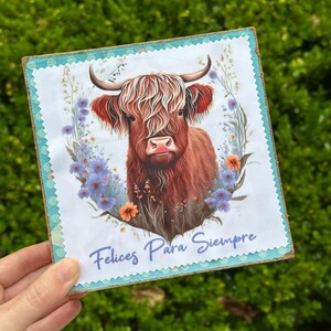 May include: Square wooden sign featuring a Highland cow illustration with long, shaggy red-brown fur, set against a floral wreath of blue and orange flowers. The sign has the text "Felices Para Siempre" in blue script.