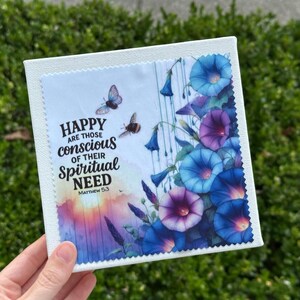 May include: A square canvas print featuring a watercolor design of blue and purple morning glory flowers, butterflies, and the text "HAPPY are those conscious of their Spiritual NEED Matthew 5:3". The print is held by a hand.