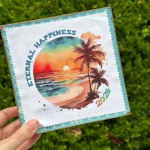 May include: Square coaster with a watercolor beach scene featuring a sunset, palm trees, and the words "ETERNAL HAPPINESS" in a circle. The year "2026" is printed on the bottom right corner. The coaster has a blue border and a natural wood edge.