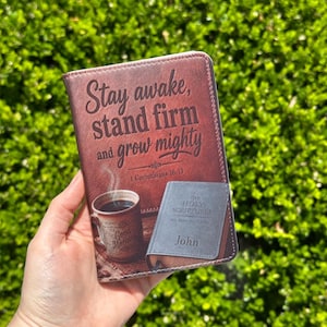May include: A rectangular brown leather-like cover featuring the printed text Stay awake, stand firm and grow mighty 1 Corinthians 16:13. The design includes a steaming coffee mug and a book titled The Holy Scriptures with the name John.