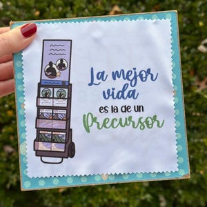 May include: A white fabric square with a blue and white illustration of a cart with photos on it. The text "La mejor vida es la de un precursor" is printed in green and black.