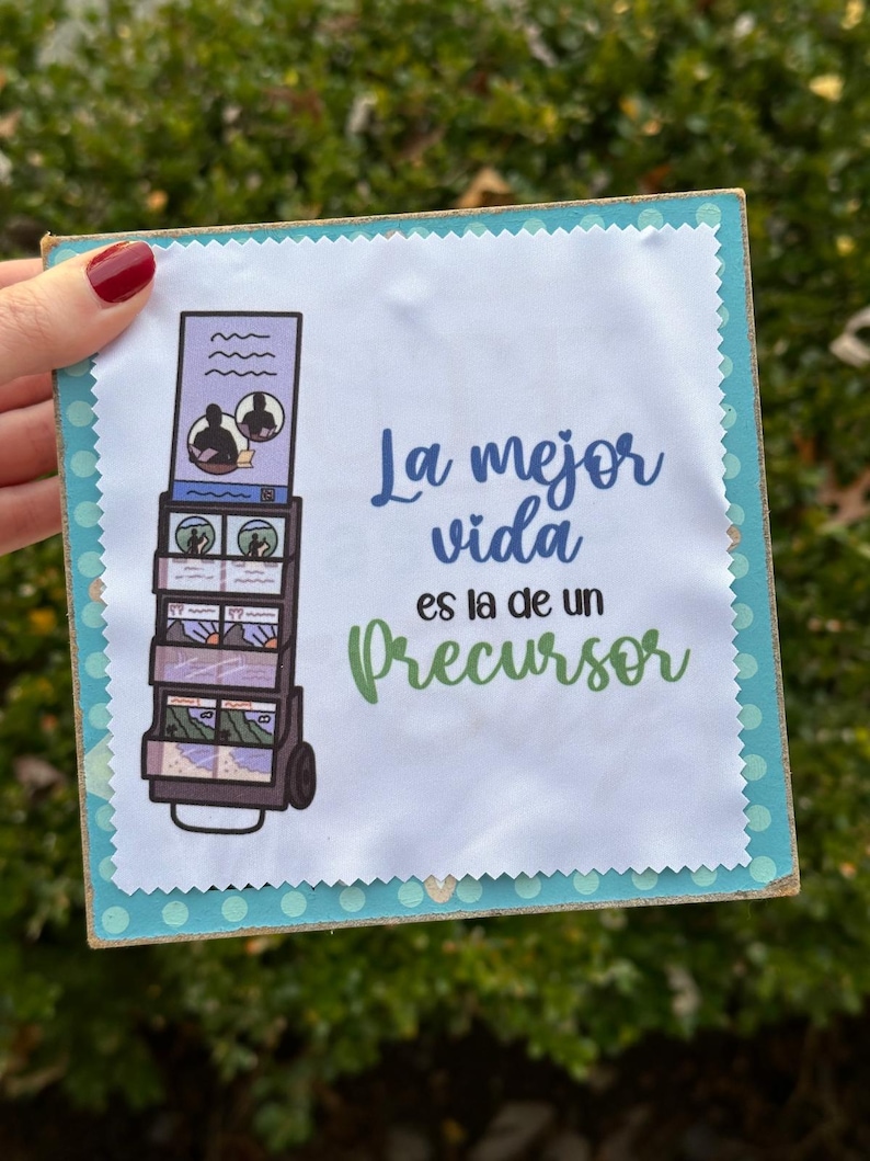 May include: A white fabric square with a blue and white illustration of a cart with photos on it. The text "La mejor vida es la de un precursor" is printed in green and black.