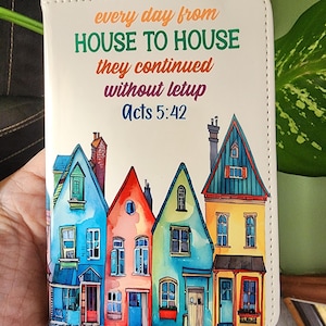 May include: A white, leather-like journal cover with colorful watercolor illustrations of houses. The cover features the text "every day from HOUSE TO HOUSE they continued without letup Acts 5:42" in orange, green, and purple.