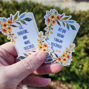 May include: Two floral stickers with inspirational quotes and bible verses. The first sticker says "Go, therefore, and make disciples" with the scripture reference "Matthew 28:19". The second sticker says "Así que vayan y hagan discípulos" with the scripture reference "Mateo 28:19".