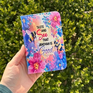 May include: A floral-themed wallet case with a vibrant design featuring pink, blue, and purple flowers, and two bumblebees. The text "TASTE and See that JEHOVAH is Good" is displayed on the front.