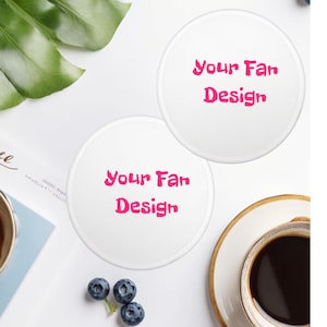 May include: Two white ceramic coasters with the text "Your Fan Design" in pink. The coasters are on a white surface, with a cup of coffee, blueberries, and green leaves in the background.