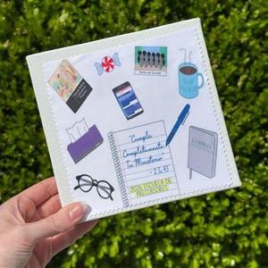 May include: A square, off-white canvas with various colorful stickers. Stickers include a coffee mug, a Bible, a notepad with text, a smartphone, a pair of glasses, and a roll of tissues. The text on the notepad reads "Cumple Completamente Tu Ministerio - 2 T 45".