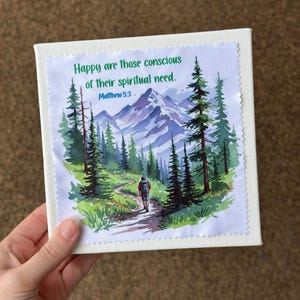 Puede incluir: Arte en lienzo cuadrado con un paisaje de acuarela que representa a un excursionista en un sendero, rodeado de &aacute;rboles y monta&ntilde;as. El texto "Happy are those conscious of their spiritual need. Matthew 5:3" est&aacute; impreso en la parte superior.