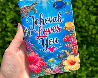 Puede incluir: Un portapasaportes rectangular con una vibrante escena submarina que muestra una tortuga marina, varios peces tropicales y arrecifes de coral coloridos. El texto "Jehovah Loves You" está impreso en una fuente estilizada con un corazón rosa.