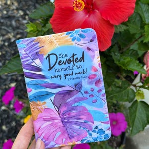 Puede incluir: Un portapasaportes rectangular con un vibrante estampado floral estilo acuarela en tonos morados, azules y naranjas. El texto "She Devoted herself to every good work! 1 Timothy 5:10" está impreso en negro en el centro de la cubierta.