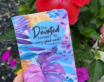 Puede incluir: Un portapasaportes rectangular con un vibrante estampado floral estilo acuarela en tonos morados, azules y naranjas. El texto "She Devoted herself to every good work! 1 Timothy 5:10" está impreso en negro en el centro de la cubierta.