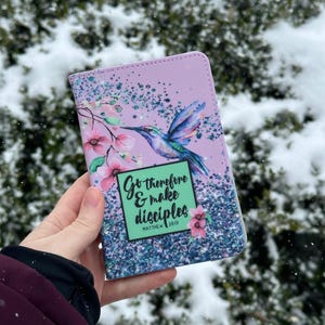 May include: A lavender-colored journal with a hummingbird design and the text "Go therefore & make disciples Matthew 28:19". The cover features a hummingbird, pink flowers, and blue accents.