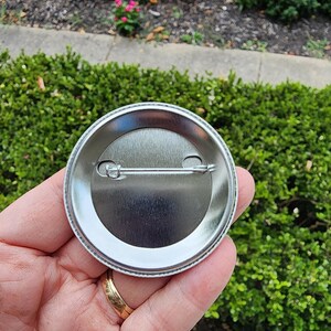 May include: The back of a silver button with a pin and a clasp. The button is round and has a smooth surface.