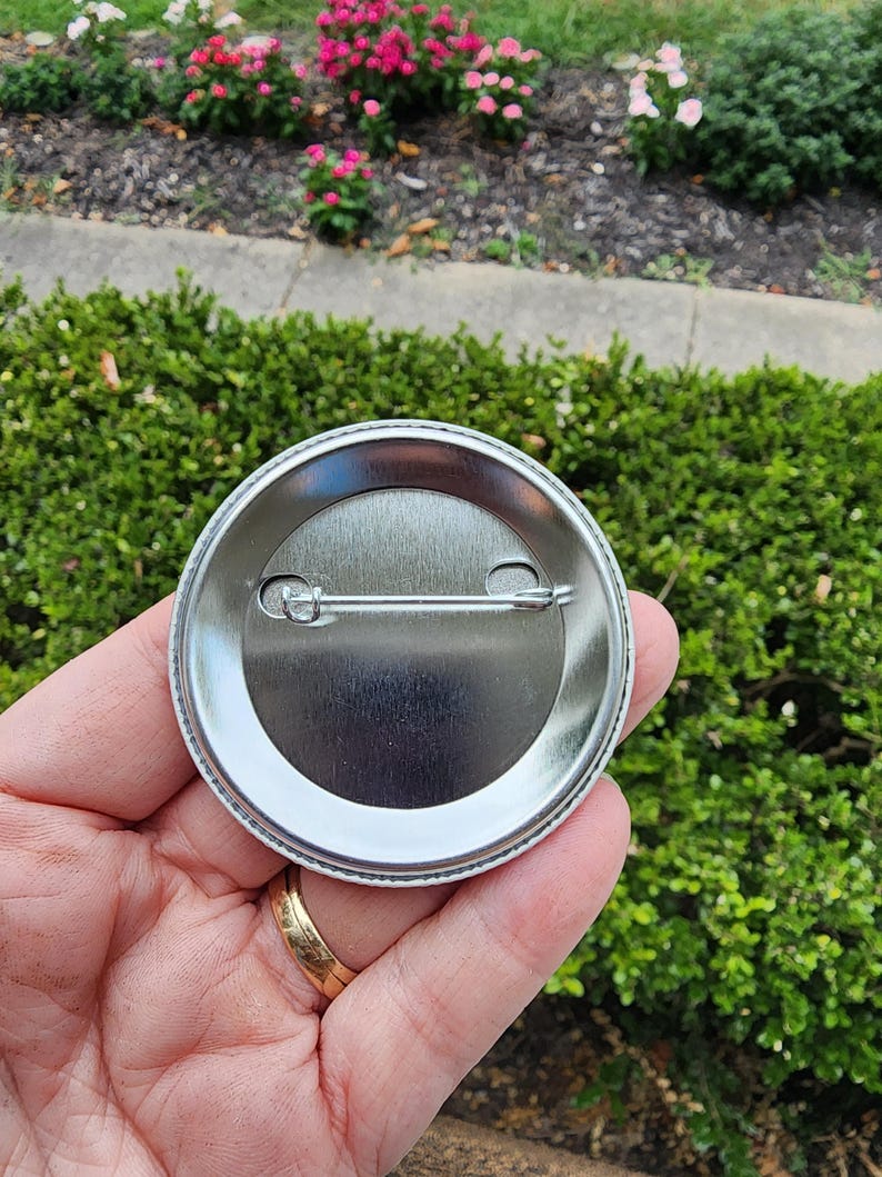 May include: The back of a silver button with a pin and a clasp. The button is round and has a smooth surface.